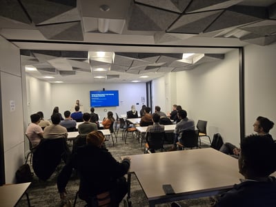 A photograph showing a presentation in a modern room with a distinctive geometric ceiling. A speaker stands at a podium next to a large screen displaying a blue slide, while an audience is seated at tables facing her.