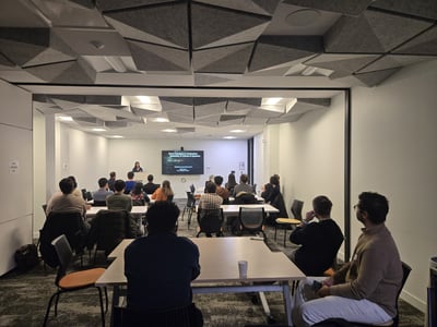 A group of people are seated in a modern conference room, attending a presentation about clinical AI systems. A female presenter stands at the front next to a screen displaying the talk's title.