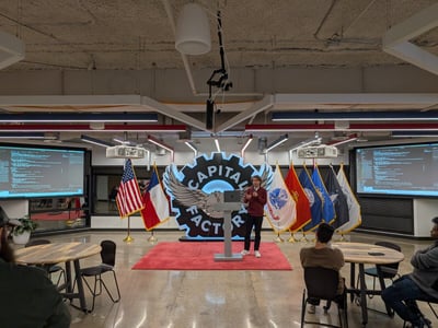 A wide-shot photograph captures an indoor presentation taking place in a large industrial-style room, where a presenter stands on a red carpet in front of a large, illuminated 'CAPITAL FACTORY' sign and a line of flags, addressing a small audience seated at round tables.