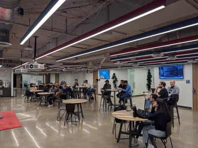 A candid photograph captures a group of approximately 20 people seated at high-top tables in a large, modern office or communal space, gathered as if attending a presentation or informal meeting.