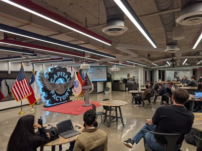 A photograph capturing an indoor presentation or event taking place in a modern office space, featuring a speaker at a podium and an audience seated at small tables facing a large, illuminated 'CAPITAL FACTORY' sign and flags.