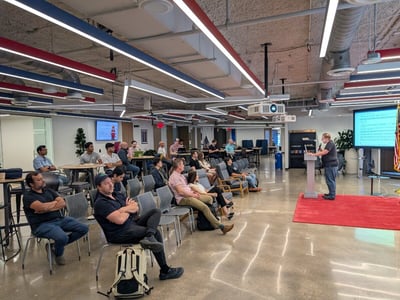 A man stands at a podium delivering a presentation to a seated audience in a modern, open-plan office space.
