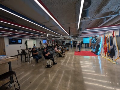 A man delivers a presentation to a seated audience in a modern, industrial-style event space featuring a large 'Capital Factory' logo and a row of military flags.
