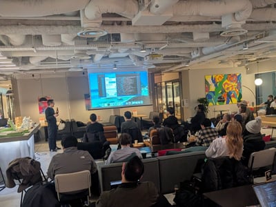 A photograph showing a technical presentation or workshop taking place indoors, where a speaker addresses a seated audience viewing code displayed on a large projection screen.