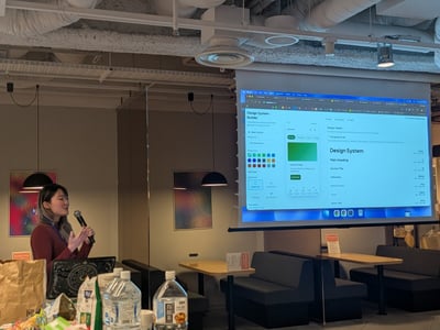 A photograph capturing a young woman giving a presentation indoors, holding a microphone, while a large projector screen behind her displays a software interface focused on a "Design System Builder."
