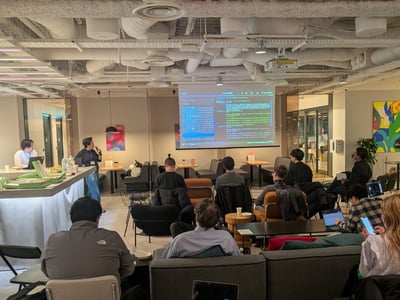 A candid photograph captures a group of people attending a technical presentation or meetup in a modern office or co-working space, watching code or terminal output displayed on a large projection screen.