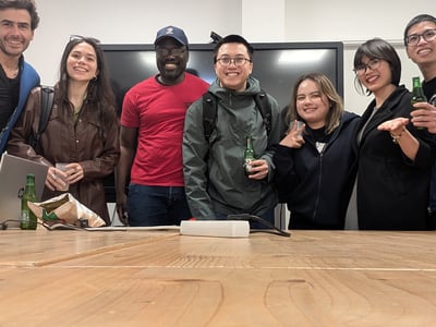 A group of seven diverse individuals are smiling and posing for a photo behind a large wooden table in an office or meeting room setting.