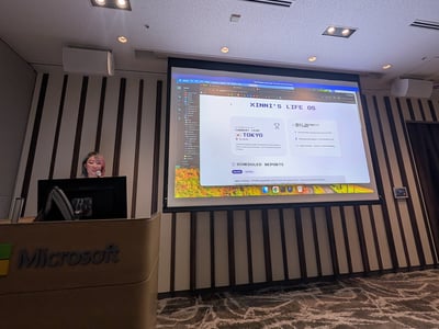A presenter stands behind a Microsoft-branded podium next to a large projection screen displaying a presentation titled 'XINNI'S LIFE OS'.