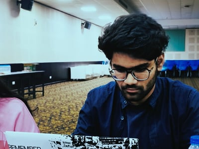 A young man with glasses and dark hair is focused on working on a laptop in a large indoor hall. The laptop's lid is covered with a black and white sticker featuring motivational phrases.