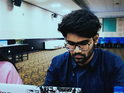 A young man with glasses and a beard is sitting at a table, focused on his laptop in a large indoor hall. The laptop is decorated with stickers that read 'REMEMBER', 'BE DIFFERENT', and 'BE YOURSELF'.