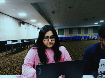 A young woman wearing glasses and a pink sweatshirt is focused on her laptop in a large, dimly lit hall.