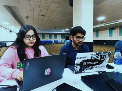 Two young individuals are seated at a white table in a large, mostly empty hall, deeply focused on their laptops.