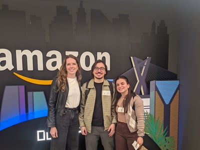 Three people stand posing together in front of a black backdrop featuring the Amazon logo and city silhouettes.