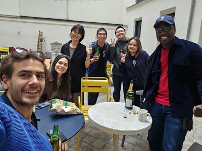 A group of seven diverse young adults are smiling and posing for a photo during a casual outdoor social gathering in a courtyard.