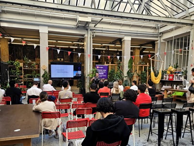 An indoor presentation taking place in a spacious, industrial-style venue with a glass skylight ceiling. An audience is seated on red chairs, facing a presenter standing next to a large display screen and a purple banner.