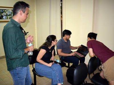 A group of four people are gathered in an indoor setting, focusing on a laptop screen held by one of them.