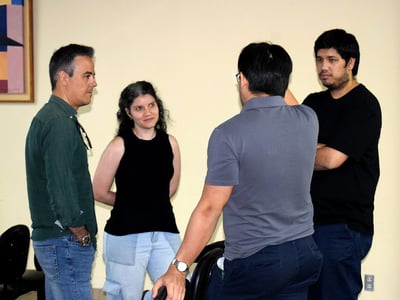 A group of four people, three men and one woman, are standing and engaged in a conversation in an indoor setting with a plain wall and a piece of art in the background.