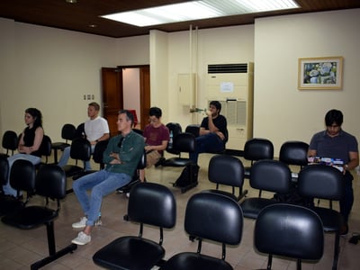 A group of people are seated in rows of black chairs within a room, appearing to attend a presentation or meeting.