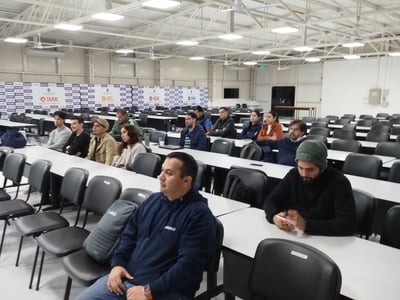 An indoor photograph of an audience seated at rows of white desks in a large lecture hall or warehouse-like classroom, listening to a presentation.