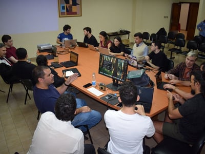 A group of people are gathered around a large wooden table in a conference room, many of them using laptops during a collaborative meeting or workshop.