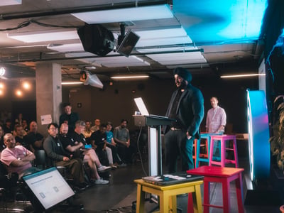 A presenter in a turban stands at a podium addressing a seated audience during an indoor event.