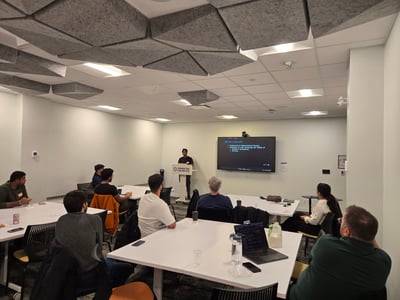 A man stands at a podium giving a presentation to a small audience seated at tables in a modern office or meeting room.