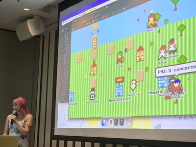 A presenter stands next to a large projection screen displaying a pixel-art style virtual simulation map.