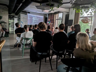 A group of people attending a technical presentation in a modern office space, with a speaker at the front and code displayed on a projector screen.