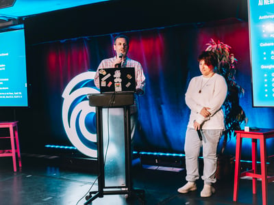 A man speaks at a podium with a laptop during a presentation, while a woman stands beside him on stage.