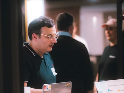 A man wearing glasses stands behind a wooden counter, looking down at a laptop decorated with stickers during an indoor event.