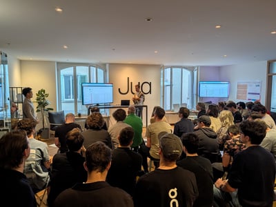 A man stands at the front of a modern office space giving a presentation to a seated audience. A large screen next to him displays a software interface, and the word 'Jua' is prominently featured on the wall behind him.