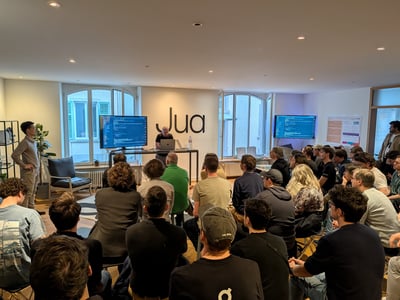 A group of people attending a technical presentation or workshop in a modern office space, with screens displaying code and a 'Jua' logo on the wall.