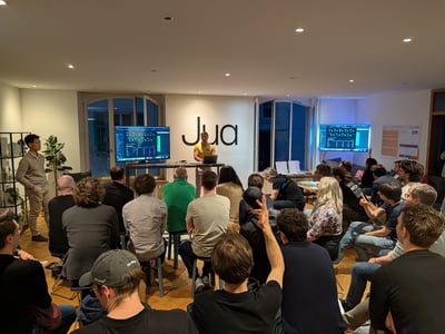 A group of people attending a tech presentation in a modern indoor space, with a presenter standing in front of a wall featuring a large 'Jua' logo.