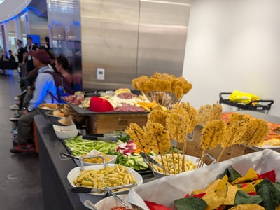 A long catering buffet table filled with various appetizers, including colorful tortilla chips, pasta salad, salsa, vegetable platters, and cheese crisps on skewers, with people sitting in the background.