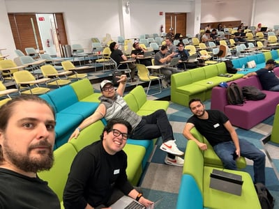 A group of people in a modern, colorful tiered workspace or lecture hall, with several men in the foreground posing for a photo while others work on laptops in the background.