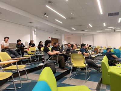 A group of people sitting in a tiered classroom or lecture hall, most of whom are using laptops and looking toward the front of the room.