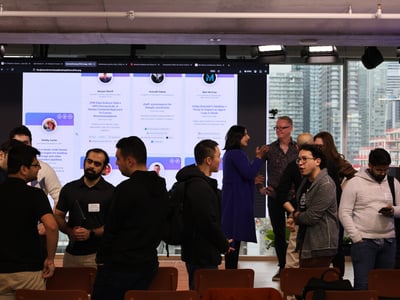 A group of people are gathered in a modern indoor space for a networking or demo event, with a large screen in the background displaying project profiles.
