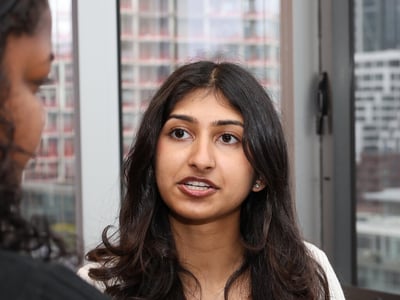 A young woman with long dark hair is engaged in a conversation, gesturing with her hand while wearing a name tag.