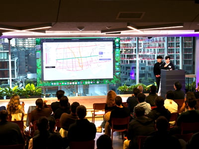 A photograph of a presentation event in a modern indoor space with a large screen displaying a map and two presenters standing at a podium before an audience.