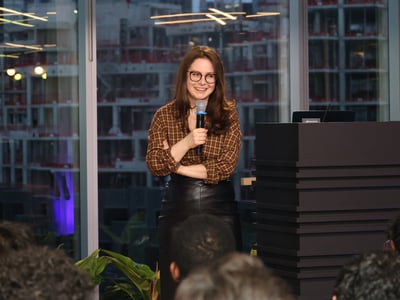 A woman with glasses and a plaid shirt stands at a podium, smiling and holding a microphone while addressing an audience.