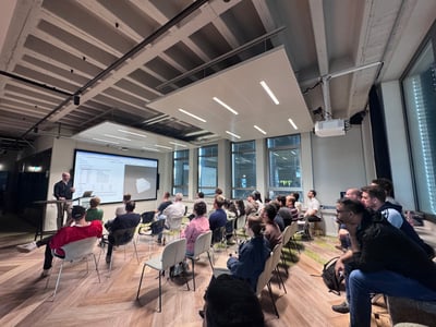 A group of people are seated in a modern indoor space, attending a presentation where a man stands next to a large screen displaying technical data and a 3D model.