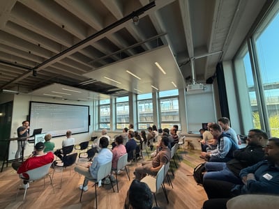 A technical presentation or workshop being held in a modern office space with an audience watching a speaker and a large screen displaying code.