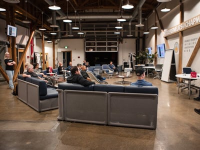 A candid photograph of an indoor event or meeting taking place in a modern, industrial-style venue, showing an audience seated on lounge sofas watching presentations on wall-mounted screens.