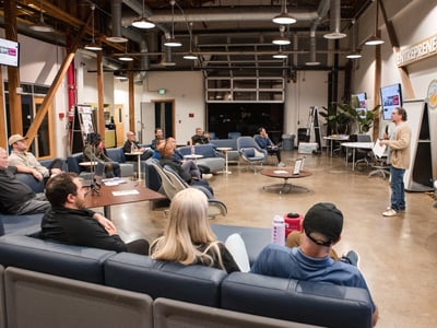 A photograph captures an evening presentation or meeting taking place in a modern, industrial-style innovation hub, where a speaker addresses a group of attendees seated comfortably on blue lounge furniture.