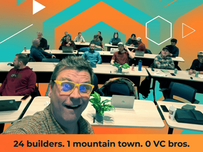 A vibrant, heavily stylized group selfie taken from a classroom or meeting, showing a man in bright yellow glasses in the foreground and approximately 15 other people seated at desks behind him, set against an orange and teal geometric background graphic.