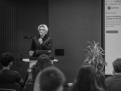 A black and white photograph of a young man with light hair giving a presentation at a podium, holding a microphone in front of an audience with a projection screen displaying a website behind him.