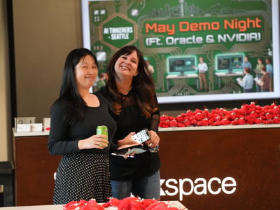 Two women smiling and posing for a photo at a tech event, with a screen in the background displaying 'May Demo Night (Ft. Oracle & NVIDIA)' and 'AI Tinkerers Seattle'.