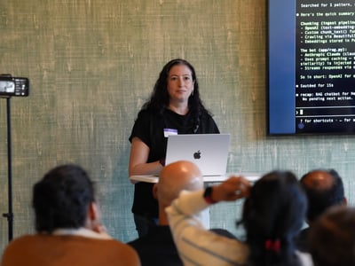 A woman stands at a podium presenting to an audience with a laptop and a large screen displaying text behind her.