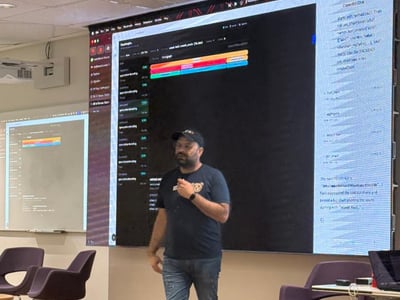 A man stands on a wooden stage, delivering a presentation in front of a large screen displaying a code editor and a chat interface.