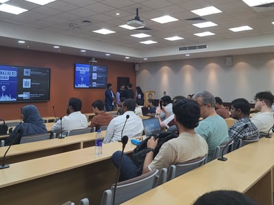 A photograph showing an audience seated in a lecture hall attending an 'AI Tinkerers Dubai' event, with presentation screens visible at the front of the room.